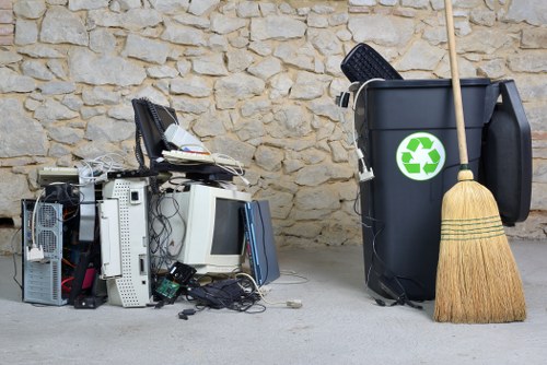 A pile of discarded electronic waste, including computer towers, keyboards, and cables, is situated on a paved surface in front of a textured stone wall. To the right, a large black rubbish bin with a green recycling symbol is positioned, partially filled with assorted waste items such as a black plastic container and some paper. An old wooden broom with a straw head leans against the bin, indicating ongoing cleaning or clearance activity. The scene appears to be part of a garden or outdoor space near Northolt, suggesting a garden clearance or rubbish removal service by Garden Clearance Northolt. The overall environment is clean, with natural daylight illuminating the debris and the surrounding area, emphasizing the need for effective waste disposal typical of garden or property clearance in the local area.
